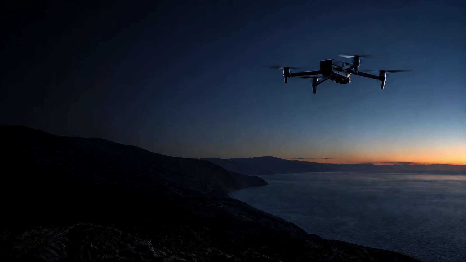 A small prosumer drone flying low along a coastal mountain ridge at dusk, the last warm horizon glow visible far in the distance
